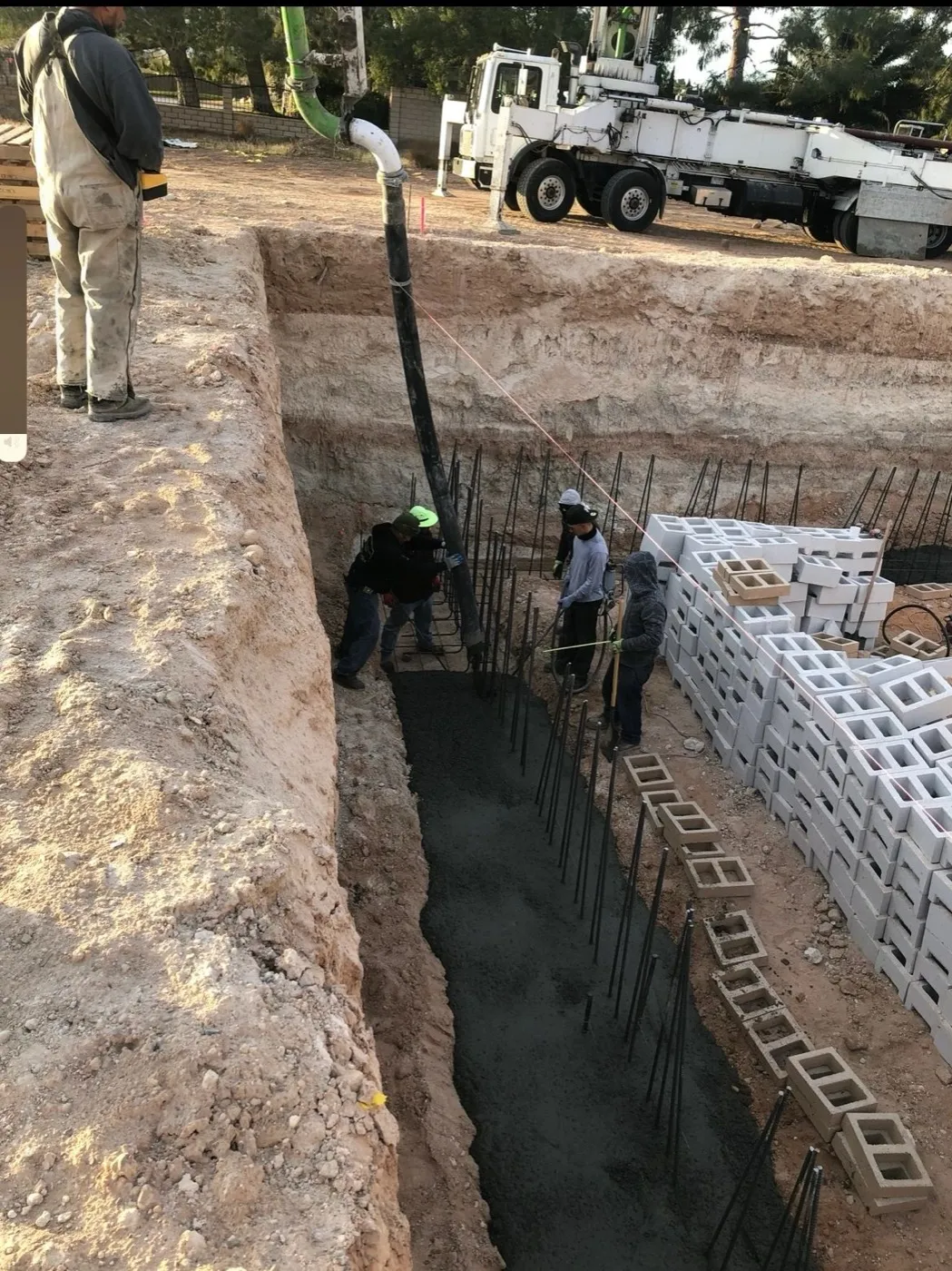 Crew placing concrete in a footing trench with the block stacked beside it for the stem wall